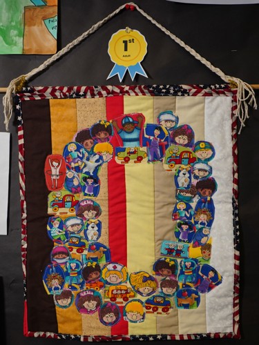 A quilted tapestry. The border of the tapestry is made with an american flag printed fabric. Stripes of fabric in skintone-like colors are layered behind quilted fabric pieces with illustrations of diverse children printed on them. The children are sticked onto the tapestry to form a ring in the center of the piece.